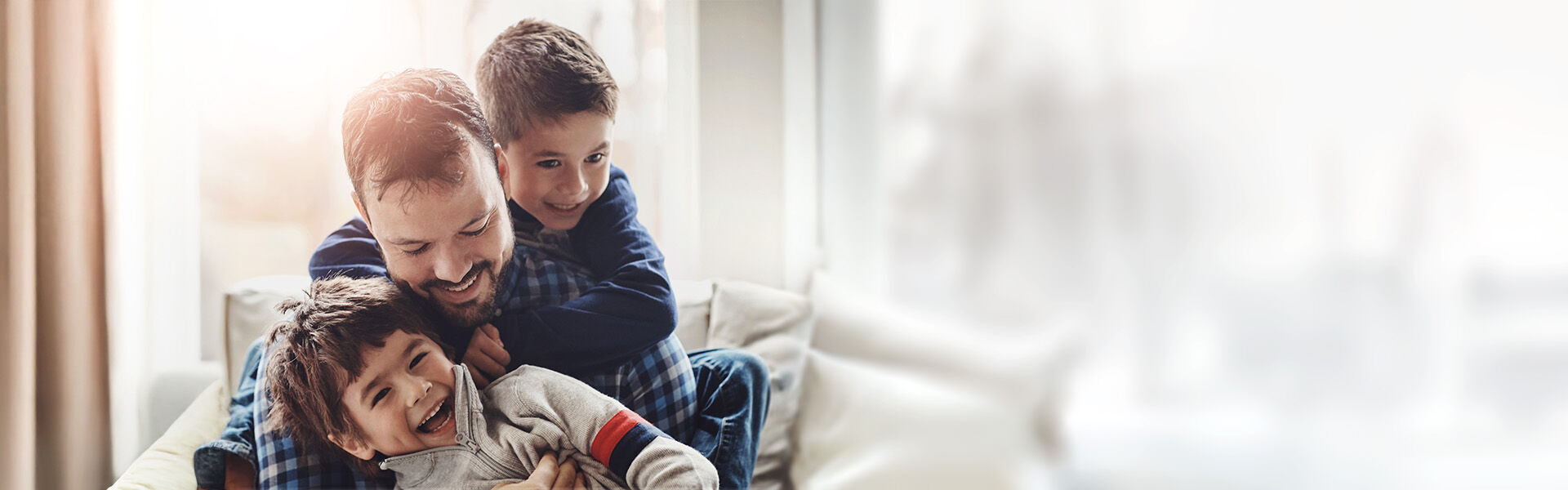 Alleinerziehender Vater sitzt im Wohnzimmer und umarmt lachend seinen einen Sohn, während der zweite Sohn ihn von hinten umarmt.