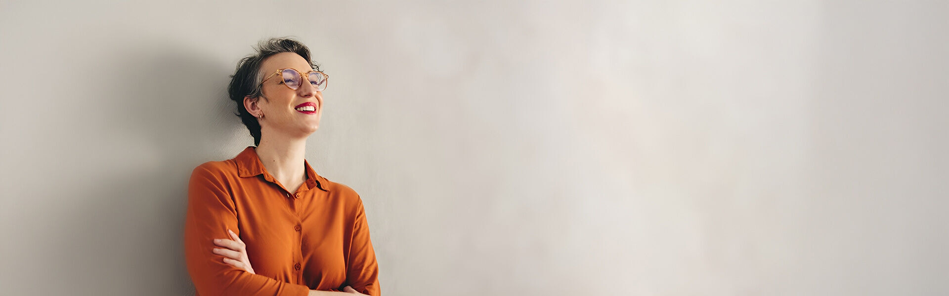 eine Frau mit orangefarbenem Hemd mit Brille lehnt an einer Wand und lächelt