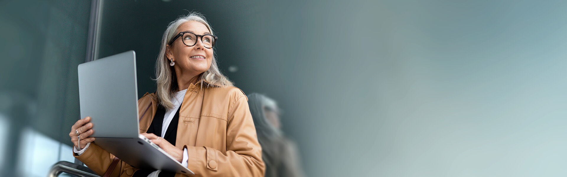 Eine Frau über 50 sitzt mit ihrem Laptop in beiden Händen und auf dem Schoß auf einem Stuhl und schaut zur Seite.