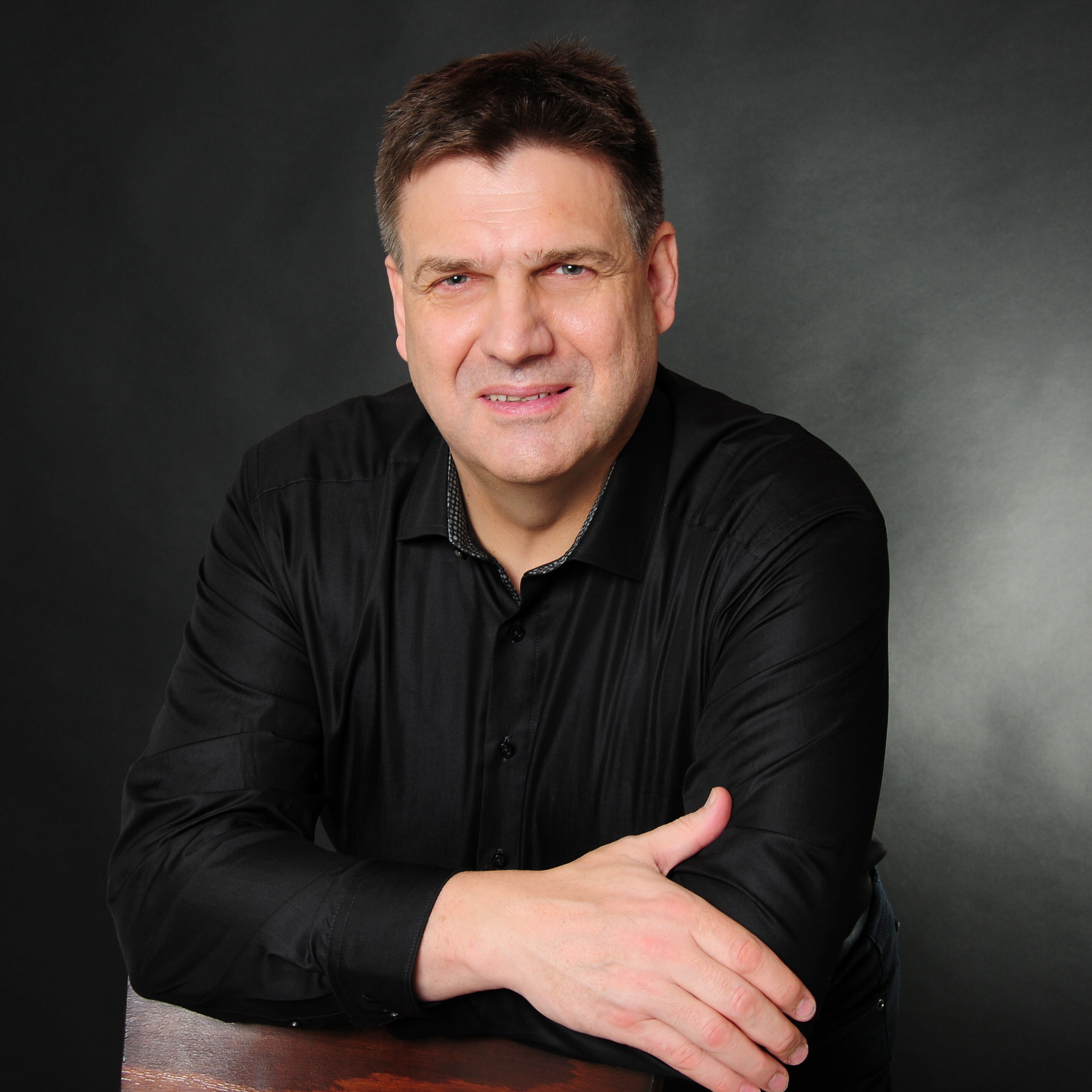 Professionelles Foto von Peter Jähne, Lernzeitbegleitung beim IBB, in dunklem Hemd vor dunklem Hintergrund.